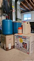 Grouped view including Koolatron cooler-warmer in original box, two large blue coolers, orange thermos, white sprayer and retro Coleman metal cooler at floor level in basement.