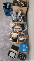 Overview photo showing blue laundry basket, cardboard box of hangers, white wire baskets, wicker basket with electronic items, boxed remote control, Kobo eReader, digital calculator.