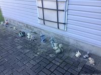 Multiple lawn figurines arranged in a line along house wall, showing variety of figures including rabbit, squirrel, angel and other animals.