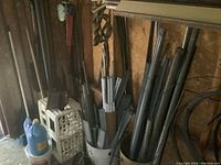 Metal pipes of varying sizes stored upright and inside a bucket along with some garden tools and a gallon jug.