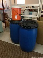 Photo of two blue plastic barrels with black lids and orange plastic pail on top, located in basement area with various household and storage items visible.
