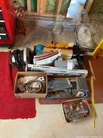 Overall view of lot showing multiple boxes containing tools, sanding paper, cords, and other accessories on floor next to red carpet.
