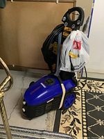 Full view of the blue Simoniz electric pressure washer with hose wrapped around the handle, white plastic bag hanging on the handle.