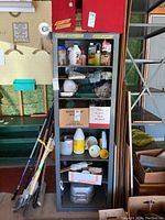 Front view of tall wood cabinet with metal frame filled with various household and cleaning items, showing shelf contents and wood grain.