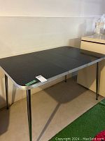 Foldable rectangular black table with four metal legs and metal edged top, photographed from corner angle.