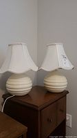 Full view of two ceramic table lamps with beehive style bases and white fabric shades with braided trim.
