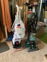 Photo of Bissell Quick Steamer Plus and Dirt Devil vacuum side by side on carpeted floor with surrounding household items.