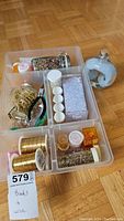 Overhead photo of the IKEA plastic organizer with multiple compartments containing various beads (clear faceted beads, multicolored seed beads), gold-colored chain, spools of gold wire, and small bead containers, plus a separate heart-shaped glass bottle filled with white beads.