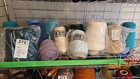 Multiple cones of yarn in different colors including teal, cream, purple, and beige arranged on metal shelving.