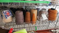 Wide view of multiple cones of yarn on metal shelving, including Bramwell Duo Magic beige cone and other cones in brown, orange, black colors
