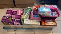 Top view showing various packets of Poise personal care liners and pads, tissue packs and a large clear plastic storage bin containing these items.