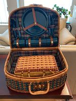 Open view of smaller picnic basket showing blue-green plastic plates, cutlery, and cups secured inside woven basket with plaid lining.