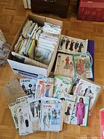 Box filled with numerous vintage sewing patterns, some organized upright and some loose, with clothing illustrations on covers showing various women's fashions.