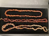 Three amber bead necklaces laid out on black velvet. Varied bead sizes and tones visible. One clasp appears broken on light-toned necklace.