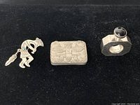 Three items on black velvet: Navajo silver Kokopelli brooch with dangling feather, engraved rectangular pill box, and Mexican silver perfume bottle with black stone top