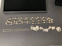 Two silver necklaces displayed on a black surface with a box in the background, showing chain and circular pendant details.