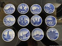 Top-down view of all 12 round blue and white Danish butter pats featuring different Danish landmarks and scenes, arranged in a grid on a dark surface.