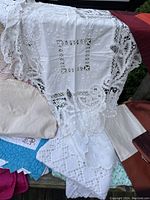 Assorted fine linens including white embroidered linen with Battenburg lace edges, crocheted lace pieces, and multiple quilts in the background.