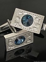 Close-up of the front of the cuff links showing the rectangular shape, blue sapphire colored round stones, and engraved silver tone metal patterns.