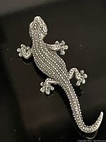 Silver-tone gecko pin shown at an angle highlighting overall shape and dot pattern texture on body and limbs.