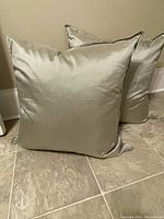 Two square pillows in smokey grey sage silk arranged on tiled floor against wall. One pillow slightly behind the other, showing smooth fabric texture and plump filling.