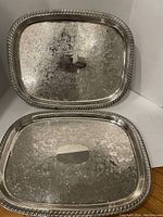 Two matching silverplate trays arranged upright showing gadroon patterned edges and faint etched design on center surface.