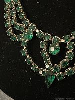 Close-up of emerald green rhinestones set in claw settings on the necklace, showing detail of the stones and metalwork.