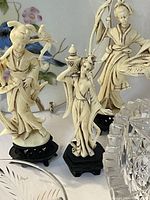 Photo of three ivory-colored Asian-inspired figurines on black stands, showing detailed features and clothing