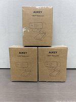 Three boxes of Aukey 1080P webcams stacked, showing brand and model on front of the boxes.