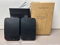 Two smart trash cans with control panels on front, one box in background. Both cans are compact and black.