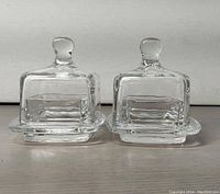 Two clear glass small square butter dishes with domed lids, front view showing the rounded knob handles on the lids.