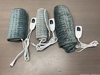 Three rolled soft flannel electric heating pads in grey, each with an attached white remote controller and power cord on a brown surface.