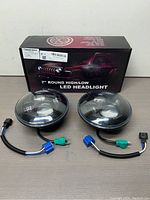 Two 7 inch round LED headlights with wiring and connectors positioned in front of the product box inside which one is partially visible. The box shows the product branding and key features.