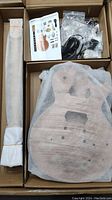 Top view of kit showing guitar neck wrapped in cloth, quilted maple top guitar body wrapped in protective fabric, small plastic bag with parts, and instruction manual with guitar image on front.