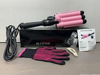 Three barrel pink hair curler, heat resistant glove, hair clip, instruction manual, and Bluetop box displayed on a gray surface.