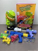 Full view of the dinosaur shooting toy set, including dinosaur blaster, foam dart guns, foam balls, safety glasses and box.