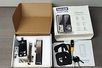 Open box showing smart lock components including rear lock unit, strike plate, and other metal parts secured in foam, with original box and user manual beside.