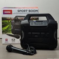 Front view of the ION Sport Boom portable Bluetooth speaker with included wired microphone and the product box in the background. Shows rugged black design and control panel.