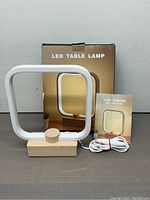 LED table lamp with square frame and wooden base, USB cable, user manual and original packaging box visible.