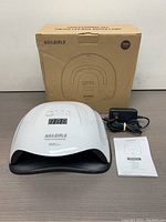 White Nailgirls Gel LED Nail Dryer Lamp with digital display and controller buttons. Shown with power adapter, instruction manual, and original cardboard box.