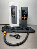 Portable tire inflator unit upright next to its box, flexible hose, USB cable and carrying pouch on table
