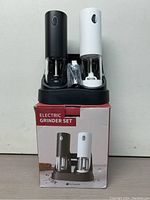 Two electric grinders (black and white) seated in their black charging base, placed on top of the product box showing the packaging design.