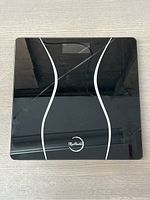 Top view of the sleek black glass smart scale showing curved white lines and digital display area on top surface.