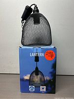 Weather-resistant outdoor lantern with black metal mesh dome, resting on its original blue packaging box marked with a price sticker.