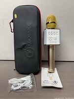 Wireless Bluetooth karaoke microphone shown upright with carrying case, charging cable, and user manual below on flat surface. Microphone has gold top and metallic gold handle with white speaker/control unit with buttons and sliders.