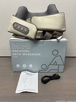 Wireless beige and gray kneading neck massager placed on its box along with user manual and power cable in front of a beige background.