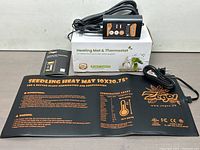 Heating mat, instructions, and thermostat with power cord displayed with box in background.