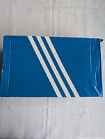 Closed blue Adidas shoe box with three white stripes on top, showing the box in good visible condition with some creasing.