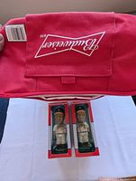 Photo showing the Budweiser logo inside the red cooler bag flap and the two unopened Oakland Athletics bobbleheads in their packages under the cooler bag.