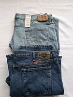 Folded view of light blue full length jeans with leather Signature Lovebrand patch on waistband and dark blue Wrangler denim shorts with leather label and red tag on waistband, showing brand and condition.